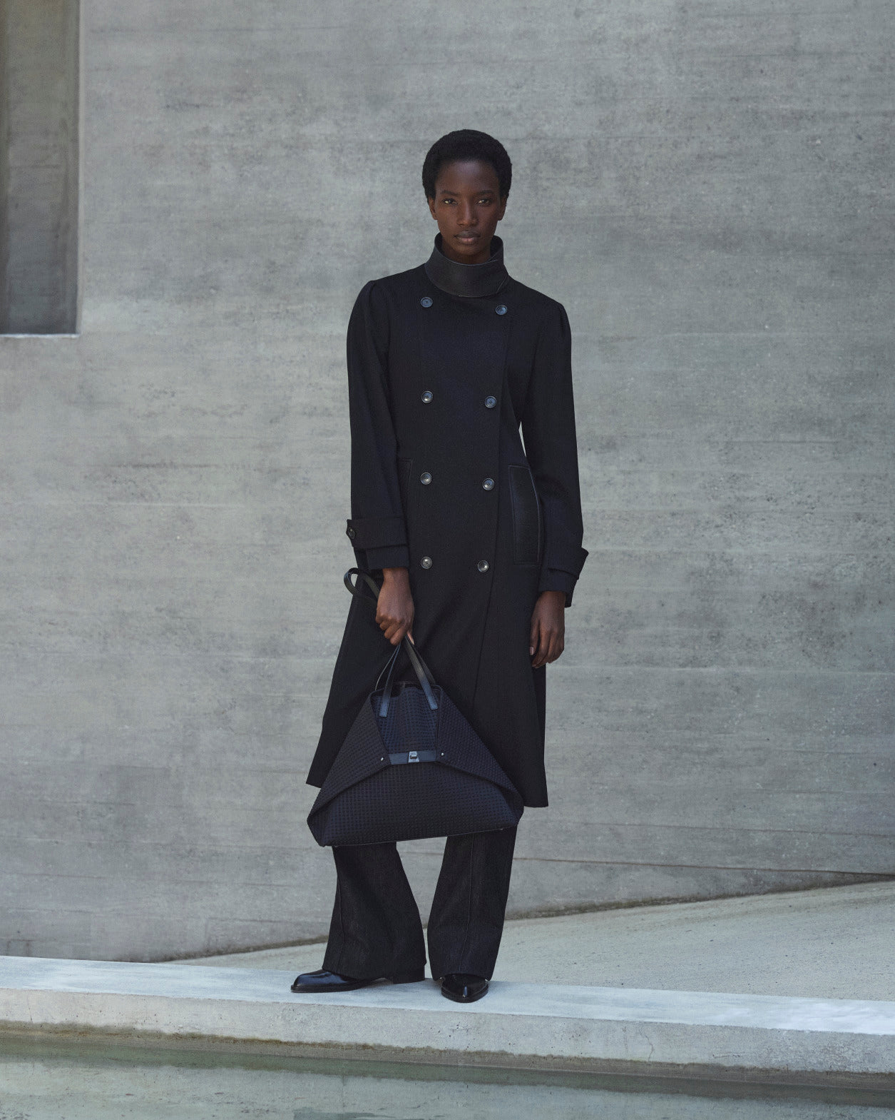 Black double-breasted midi coat styled on a female model with tailored trousers and structured leather handbag.