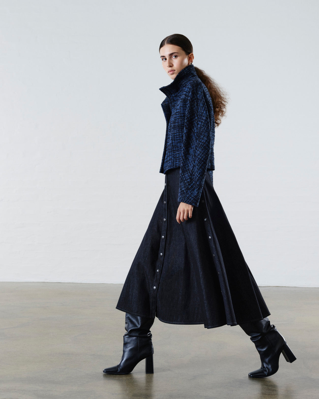 Luxury womenswear look featuring a textured blue cropped jacket, black flared midi skirt and black heeled boots, styled on a female model in a minimalist studio setting.