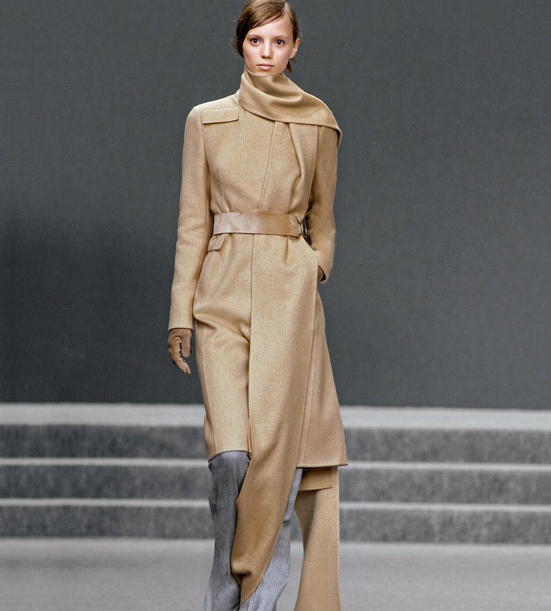 Model on the runway wearing a long beige coat and a long scarf in the same colour.