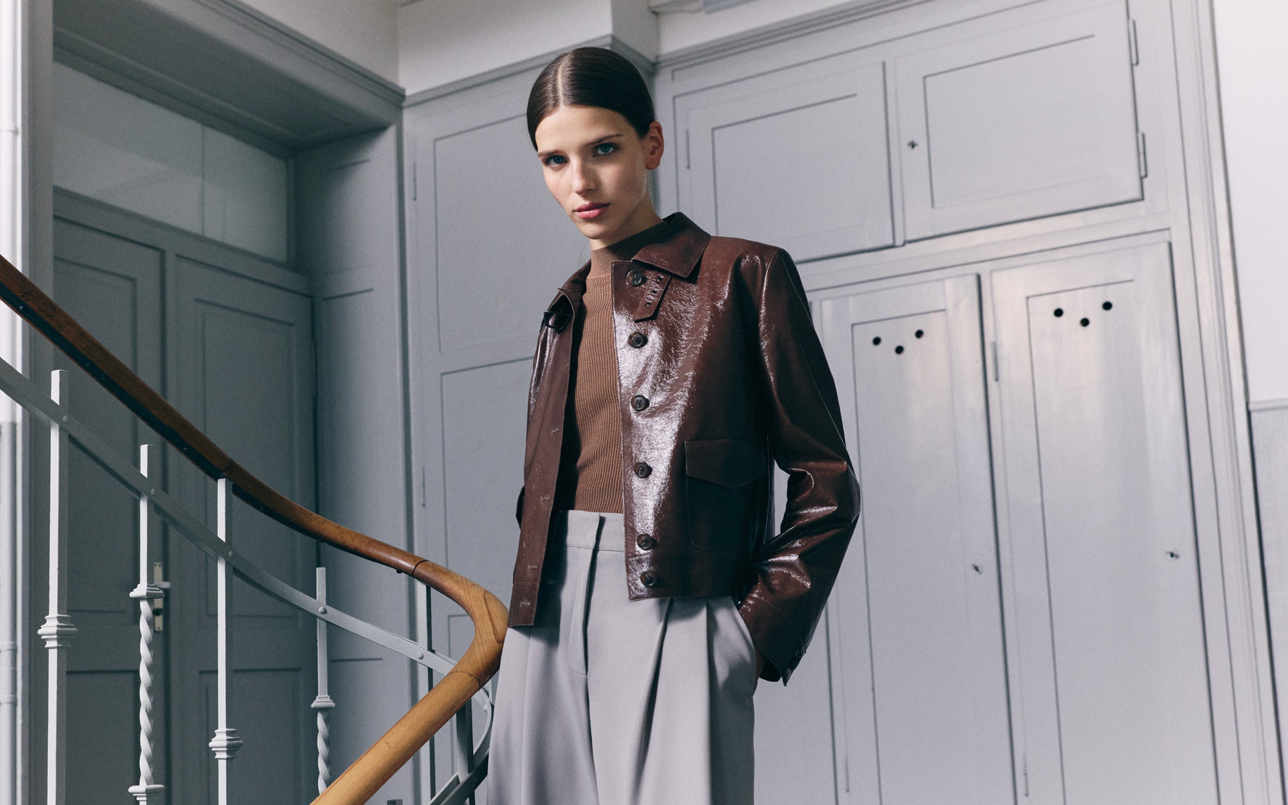 A model poses in a minimalist interior for the Akris Cruise 2026 collection, wearing a glossy chocolate brown patent leather jacket over a brown knit top and high-waisted grey pleated trousers.