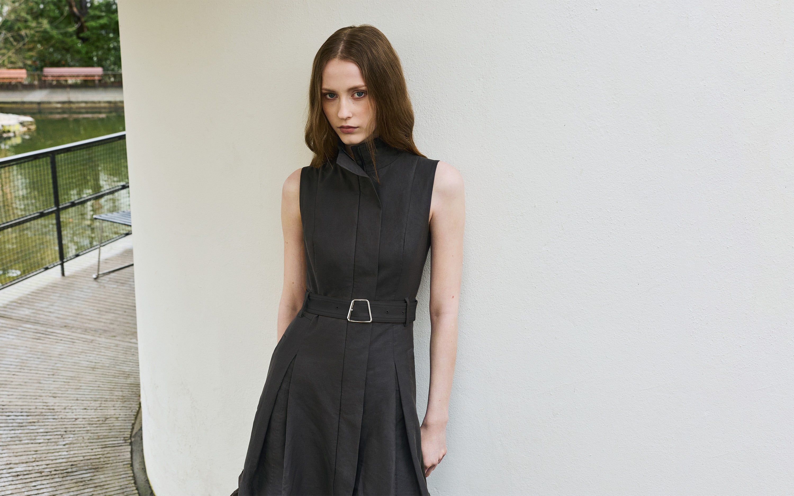 Akris Spring 2026 collection: A model wearing a sleeveless dark grey or black structured dress with a high collar and a belted waist, leaning against a white wall.