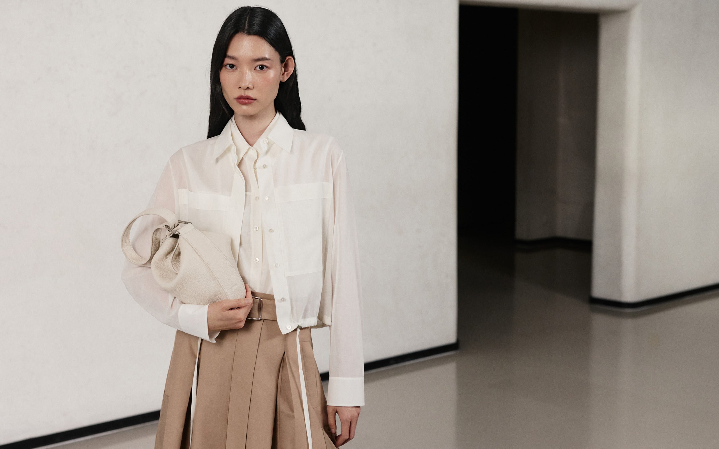 Model wearing a white silk shirt, beige skirt, and an ivory leather bag from the Akris Pre-Fall 2026 collection.
