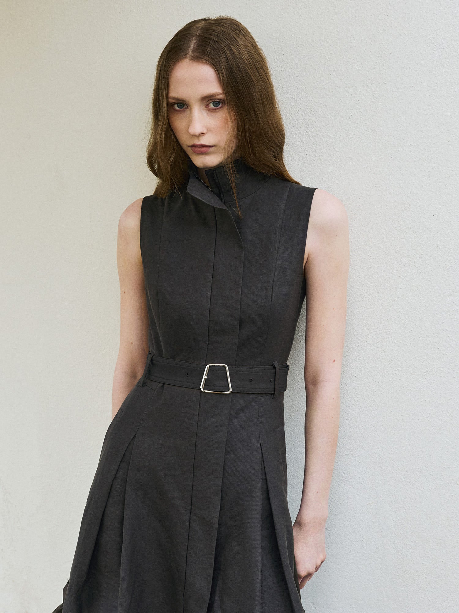 Akris Spring 2026 collection: A model wearing a sleeveless dark grey or black structured dress with a high collar and a belted waist, leaning against a white wall.