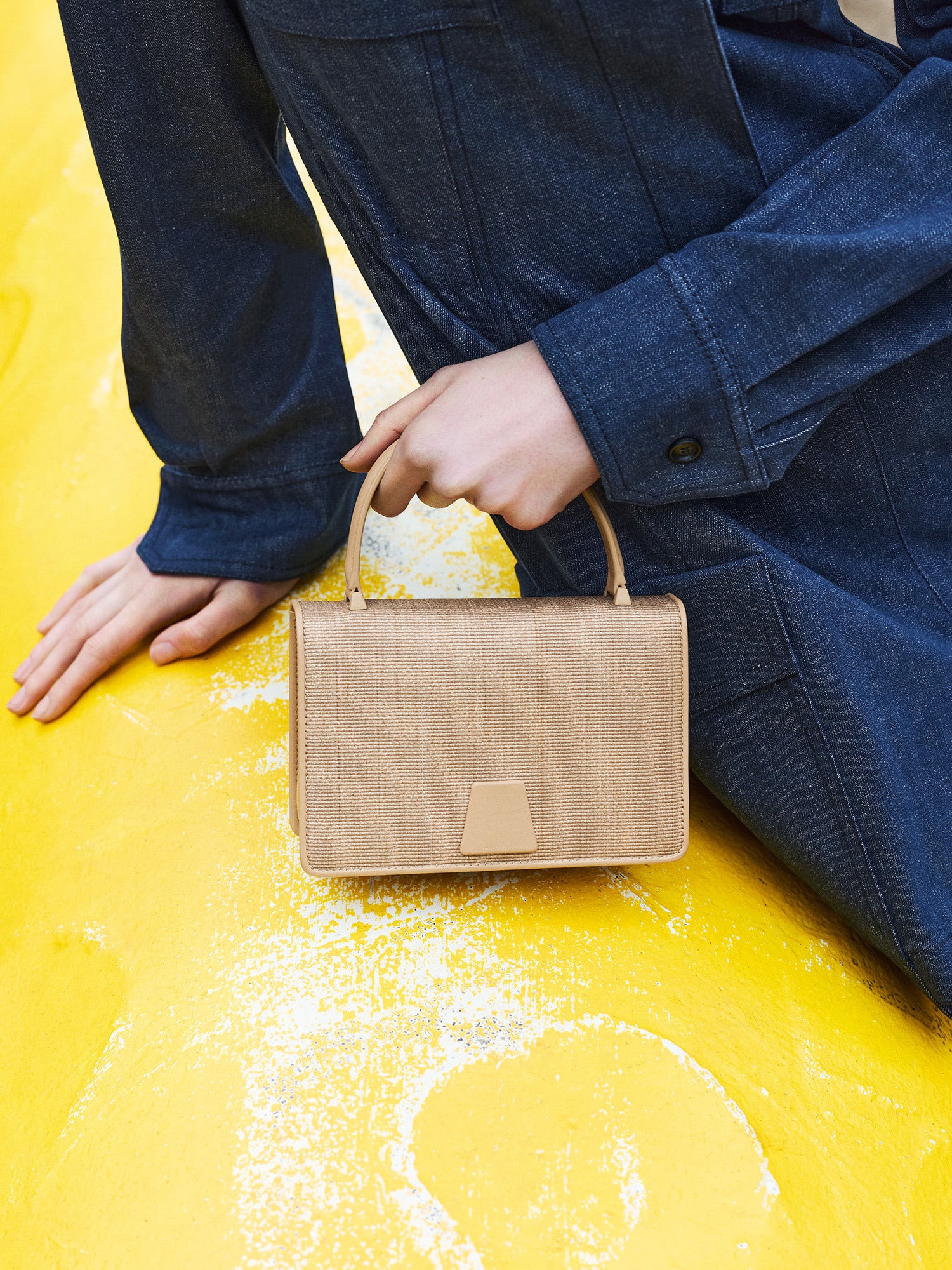 A model wears an Akris dark blue denim jacket and sits on a yellow surface with distressed paint, holding an Akris Ai bag. The bag is made of natural woven straw, featuring a single, textured light brown leather top handle and a secure trapezoidal flap closure in the same leather, positioned center-front.