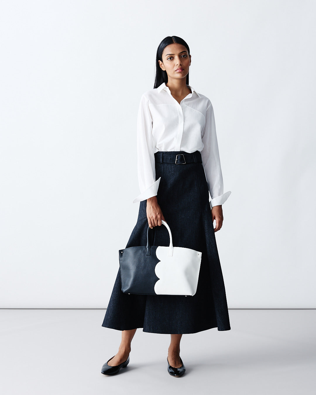 Detailed product shot of a white button-down shirt paired with a dark navy belted A-line denim skirt. The look features a structured black and white color-block tote bag.