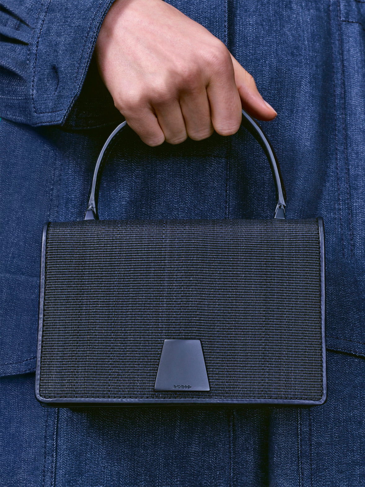 Close-up of two textured handbags with a trapezoidal clasp, one in vibrant red and the other in sleek black.
The bags are held against matching red and dark denim outfits, showcasing their fine woven detail.