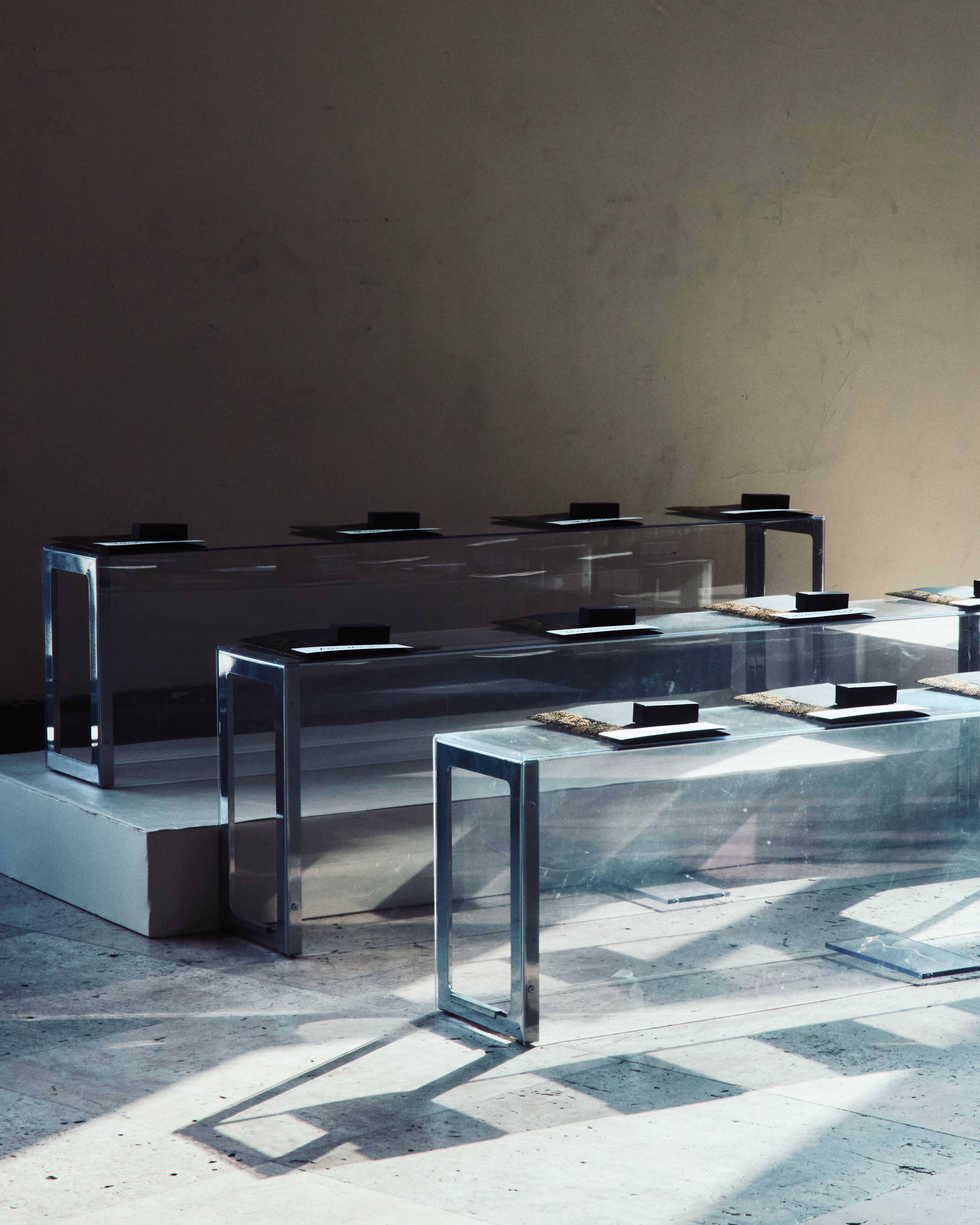 A minimalist view of transparent plexiglass benches arranged in a studio, reflecting natural light and holding small black gift boxes for guests.