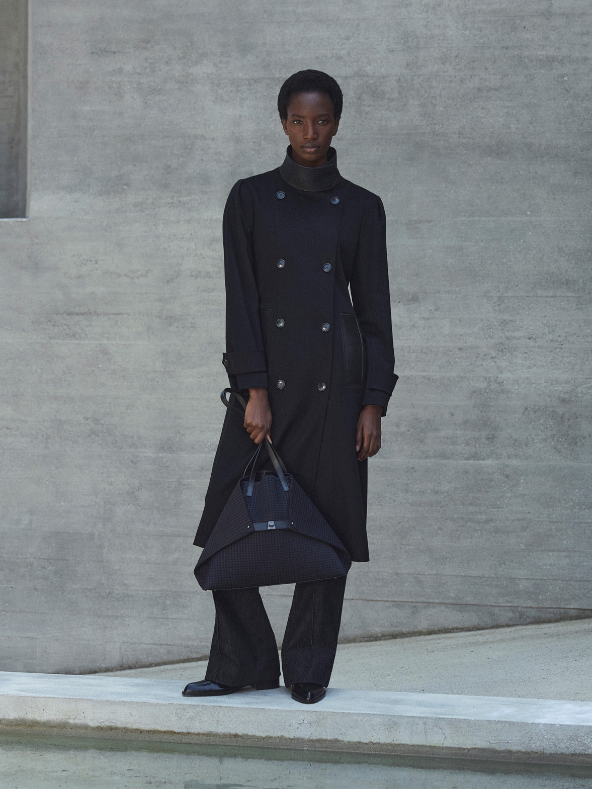 Split view of a model wearing a long black double-breasted wool coat and carrying the signature Akris Ai trapezoid bag in black textured fabric.