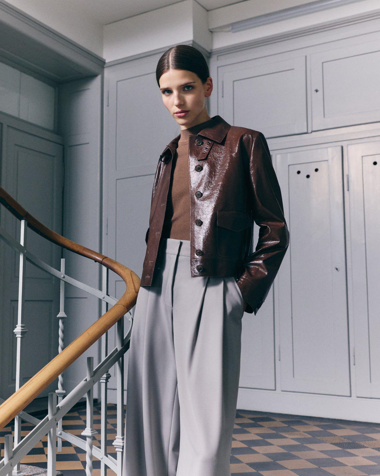 A model poses in a minimalist interior for the Akris Cruise 2026 collection, wearing a glossy chocolate brown patent leather jacket over a brown knit top and high-waisted grey pleated trousers.