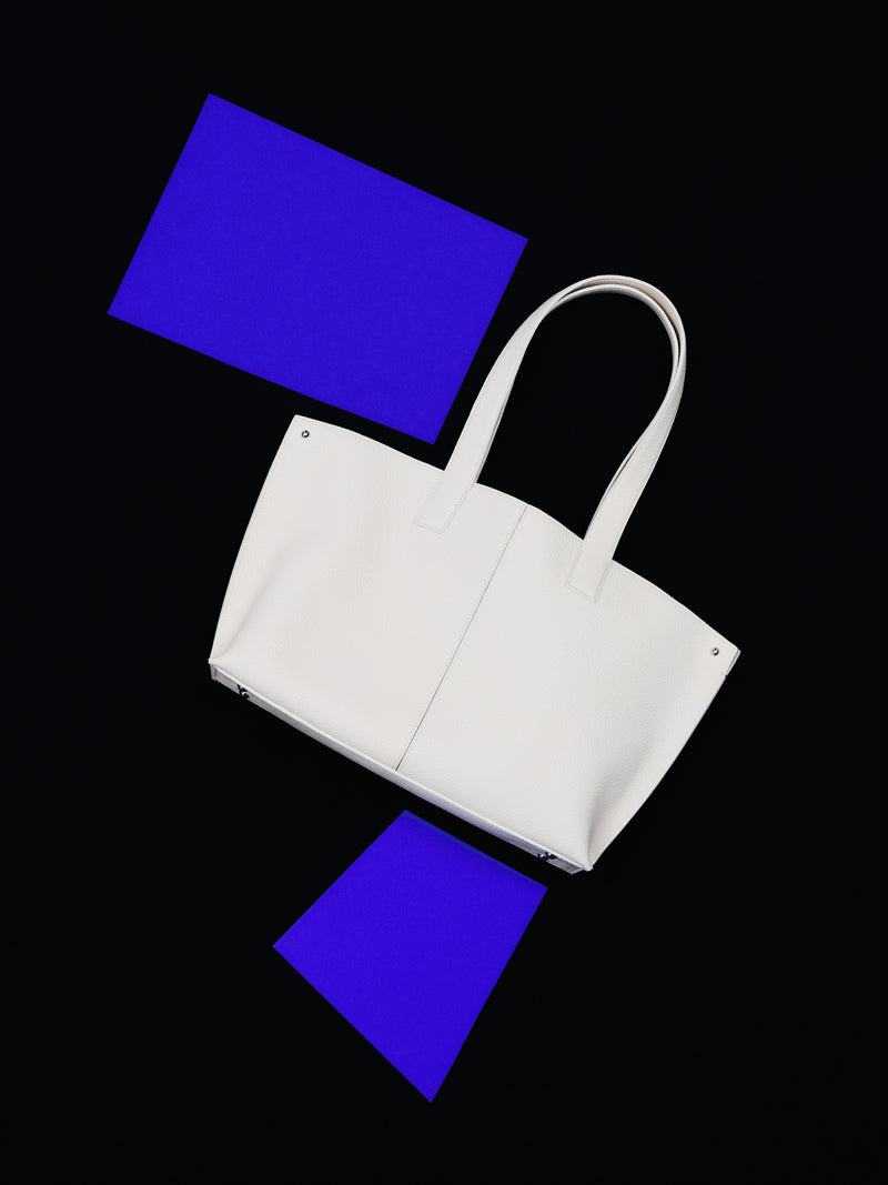 Discover the minimalist perfection of the Akris Pre-Fall collection, highlighting the iconic Ai tote bag in a pristine white leather finish. Artfully arranged against a dramatic black backdrop with vivid electric blue geometric accents, this luxury accessory embodies pure modern elegance. Elevate your collection with timeless Swiss design.