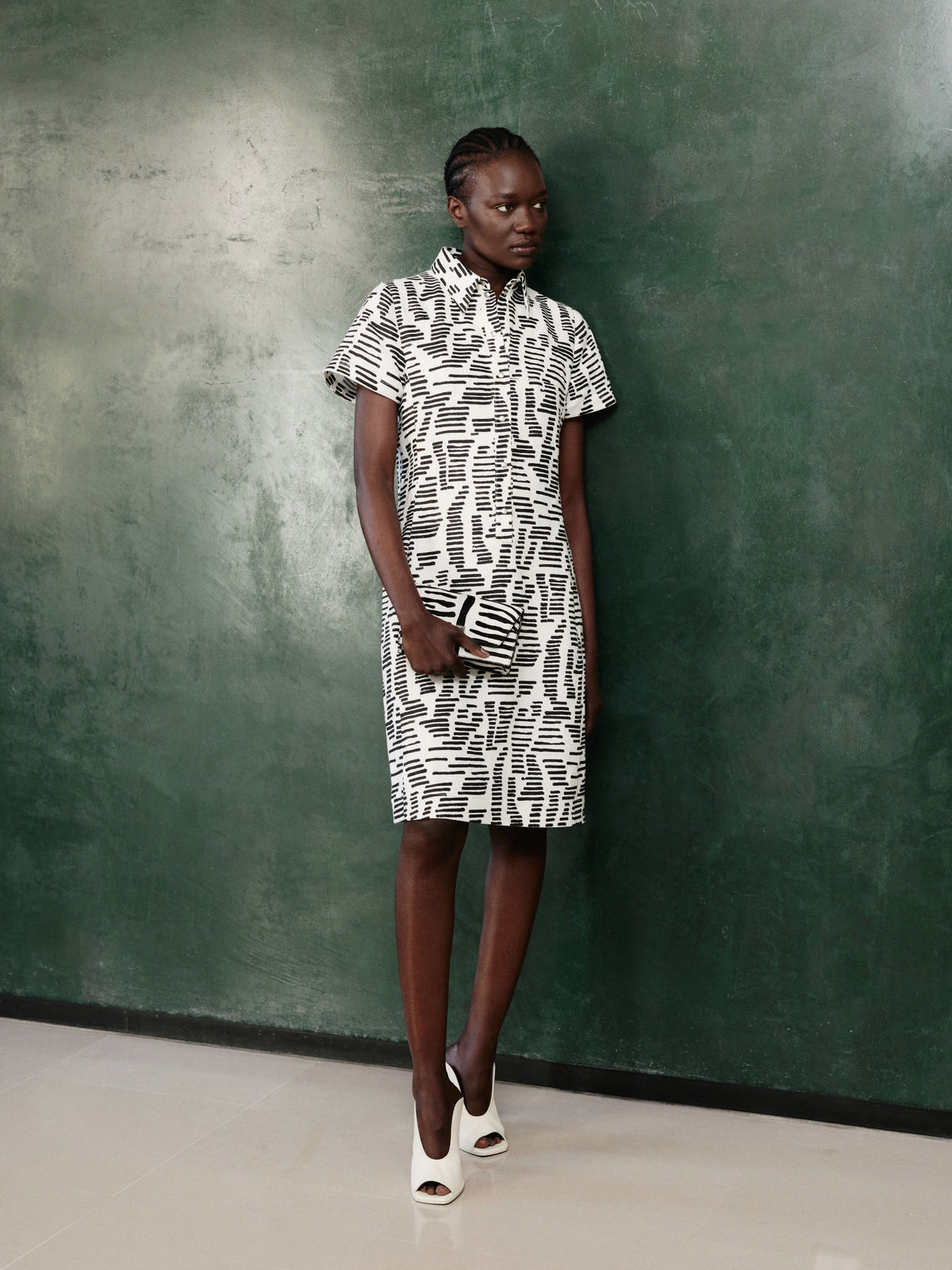 Discover the graphic elegance of the Akris Pre-Fall collection, highlighting a classic shirt dress featuring an abstract black-and-white print. The sophisticated look is paired with a matching luxury clutch bag and white open-toe mules. Elevate your everyday style with effortless modern design and premium Swiss craftsmanship.