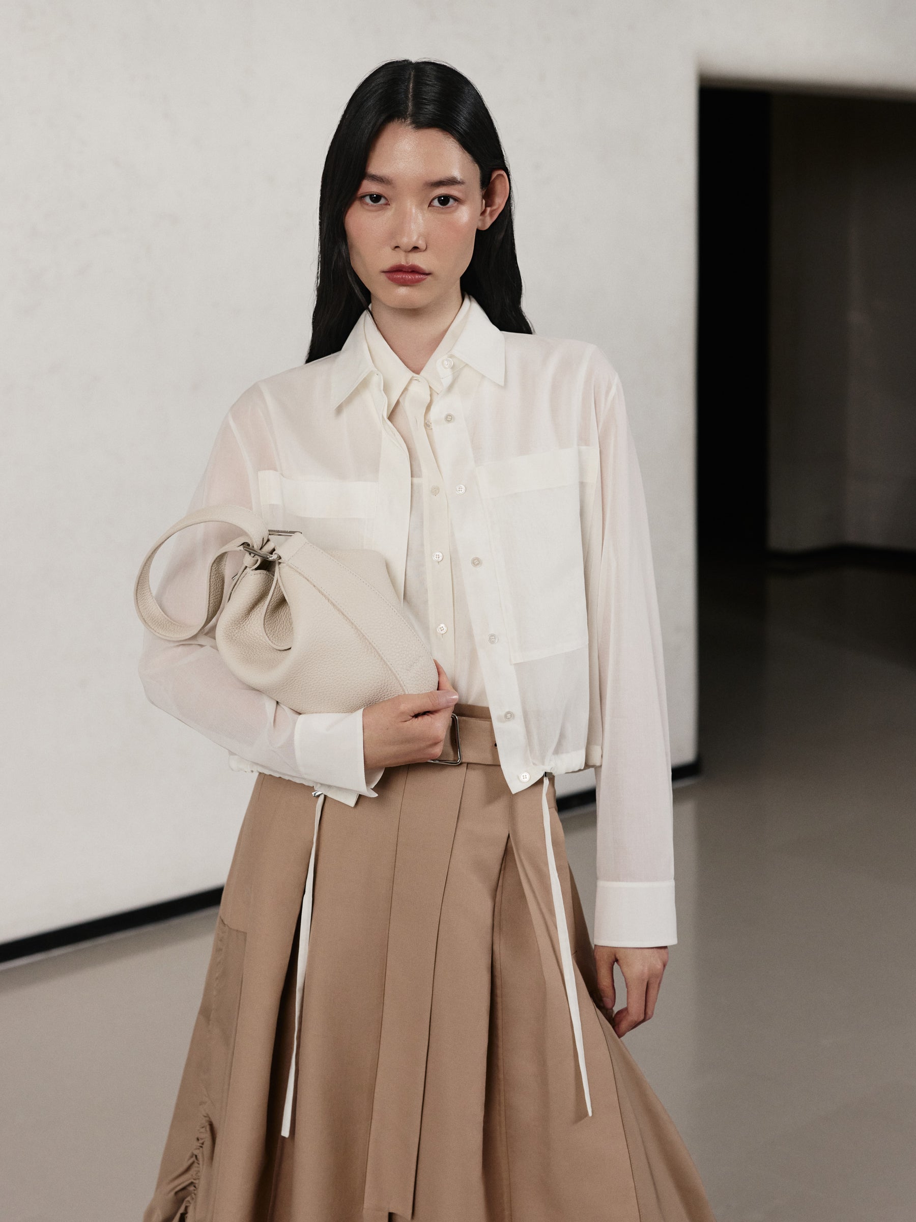 Embrace effortless refinement with the Akris Pre-Fall collection, showcasing a classic white shirt paired with a tailored beige pleated skirt. The look is beautifully elevated by the structured yet slouchy Anna Hobo bag in a soft off-white textured leather. Discover timeless, minimalist luxury and exceptional Swiss craftsmanship.