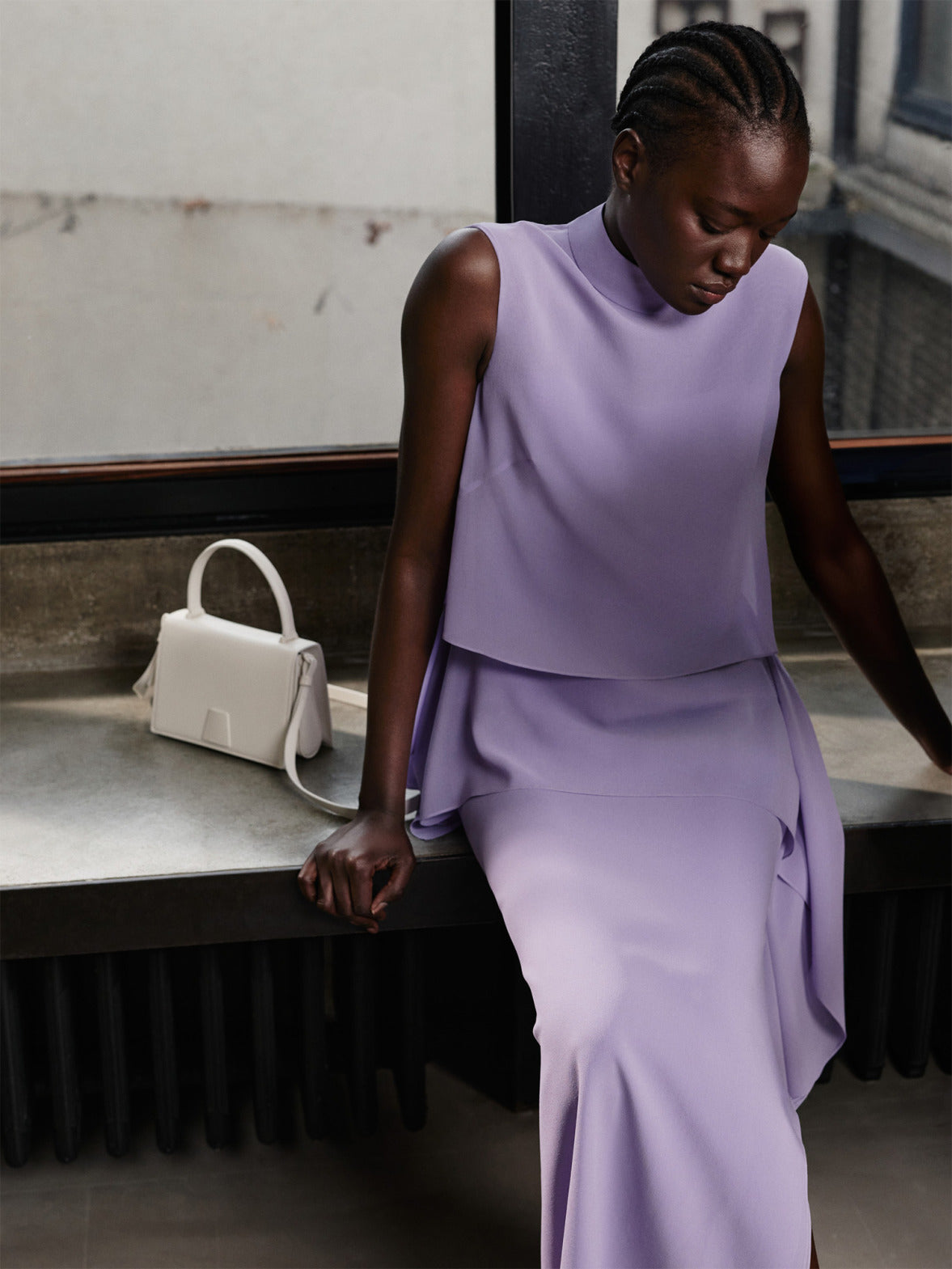 Model styling a sleeveless lilac top and matching skirt featuring lavender, the color of the season, from Akris Pre-Fall 2026. Paired with a structured white leather handbag for a modern, elegant luxury look.