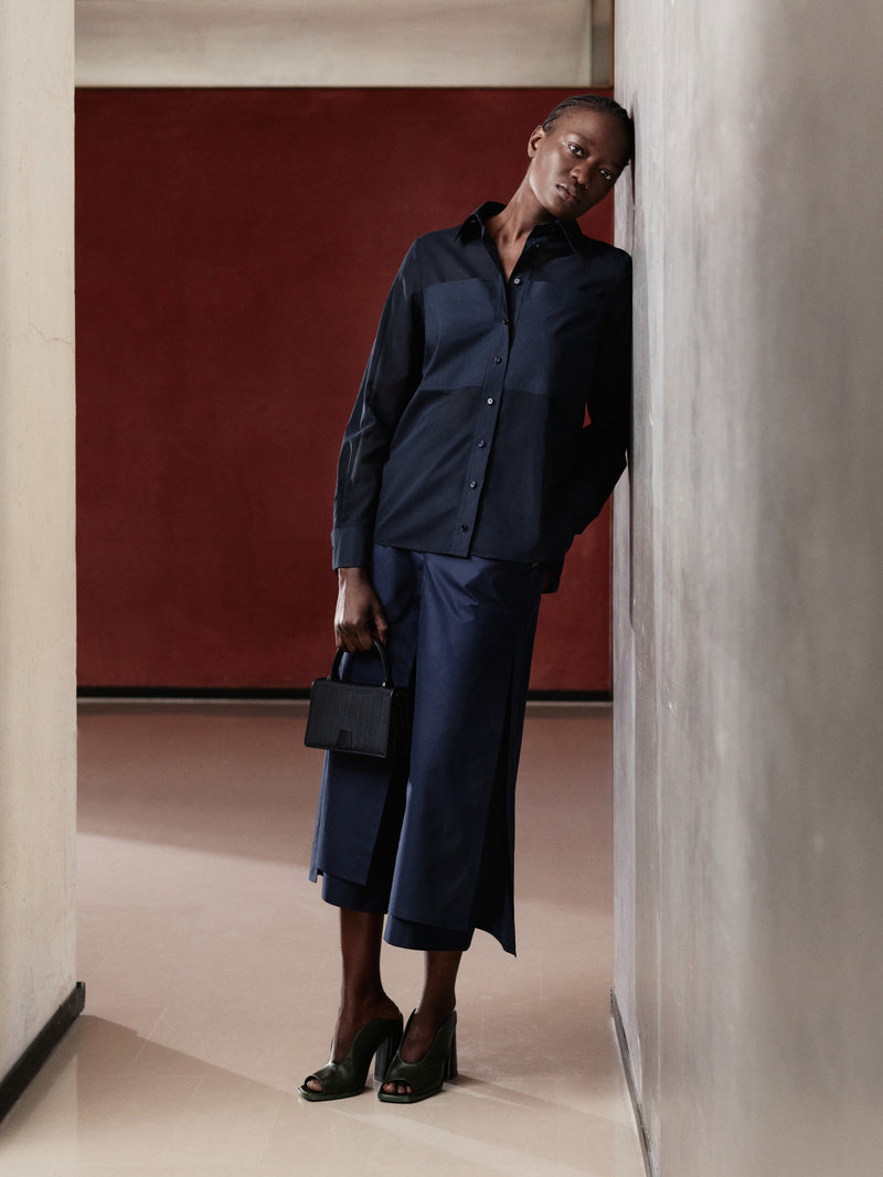 Discover the refined minimalism of the Akris Pre-Fall collection, showcasing a sophisticated monochromatic look in deep navy blue. This elegant shirt and wide-leg trousers ensemble is perfectly completed by a structured black luxury handbag. Elevate your wardrobe with fluid tailoring and timeless Swiss craftsmanship.