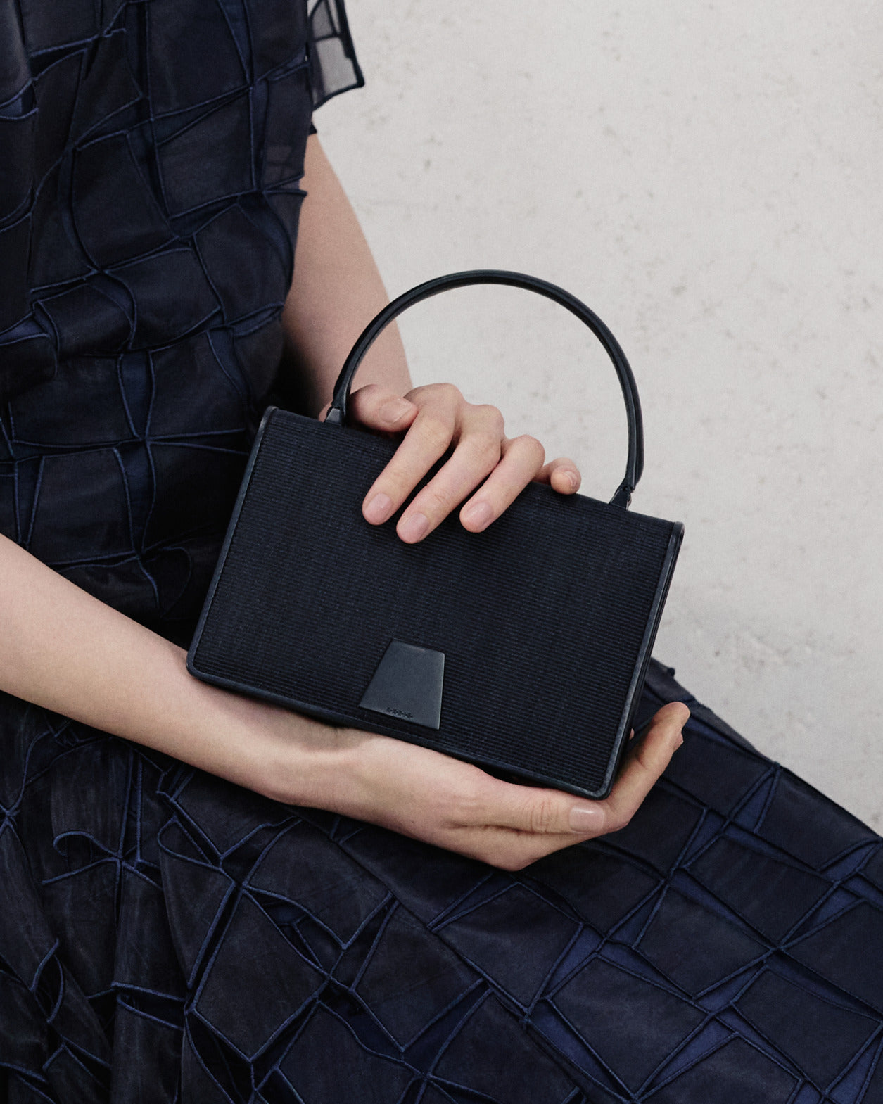 Akris Pre-Fall 2026 collection luxury black textured handbag.
Elegant structured designer purse featuring a sleek top handle and signature clasp.
Paired with a dark navy blue geometric patterned dress.