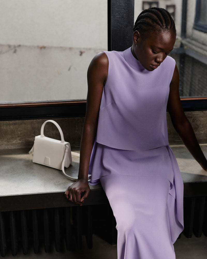 Akris Pre-Fall 2026 luxury fashion collection showcasing a lavender sleeveless top with a high neckline.
The matching draped lavender skirt creates an elegant monochrome ensemble.
Accompanied by a structured off-white leather handbag with a top handle and shoulder strap.