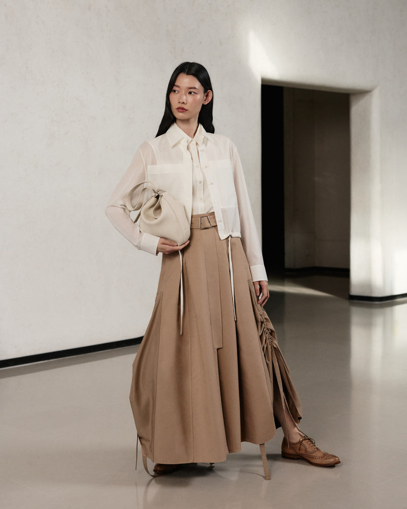 Akris Pre-Fall 2026 collection luxury coordinates for women.
White sheer long-sleeve button-up blouse paired with a belted beige maxi skirt.
Features a matching cream leather handbag and classic brown brogue shoes.