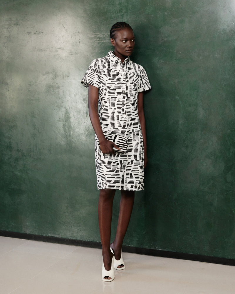 Akris Pre-Fall 2026 collection shirt dress and matching clutch bag.
Features a modern black and white graphic pattern with abstract strokes.
Short-sleeved designer knee-length dress styled with open-toe mules.