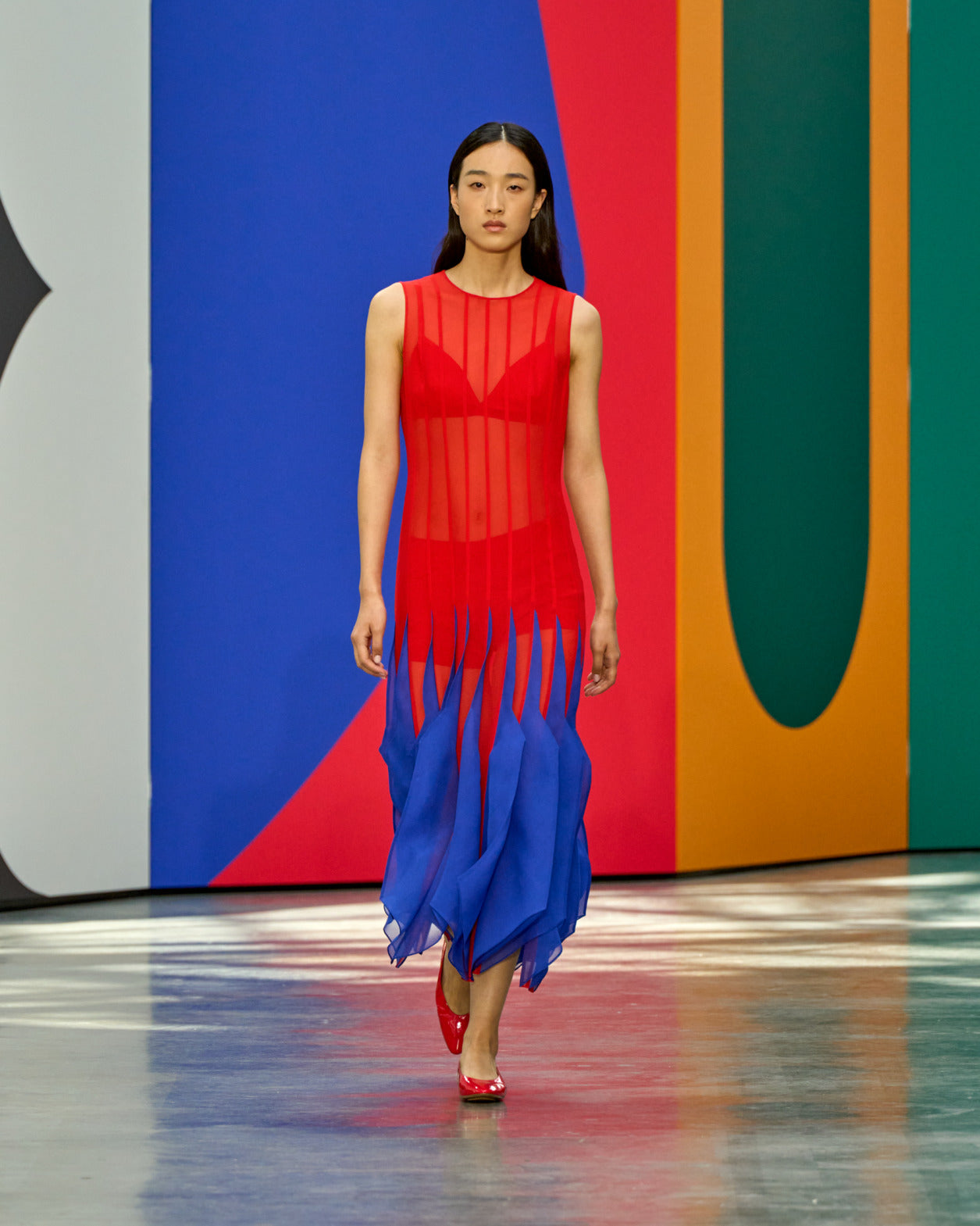 A model walks the runway in a sleeveless red sheer dress with a blue fringe and layered skirt hem, paired with red heels, against a bold multicolour geometric backdrop.