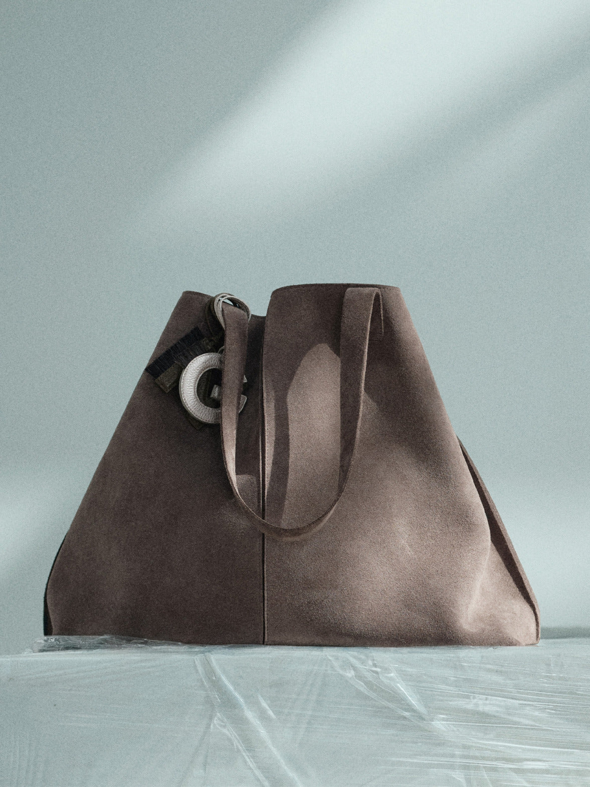 Minimalist detail shot of a woman in an olive green suede suit holding a large brown suede tote bag against a grey gradient background.