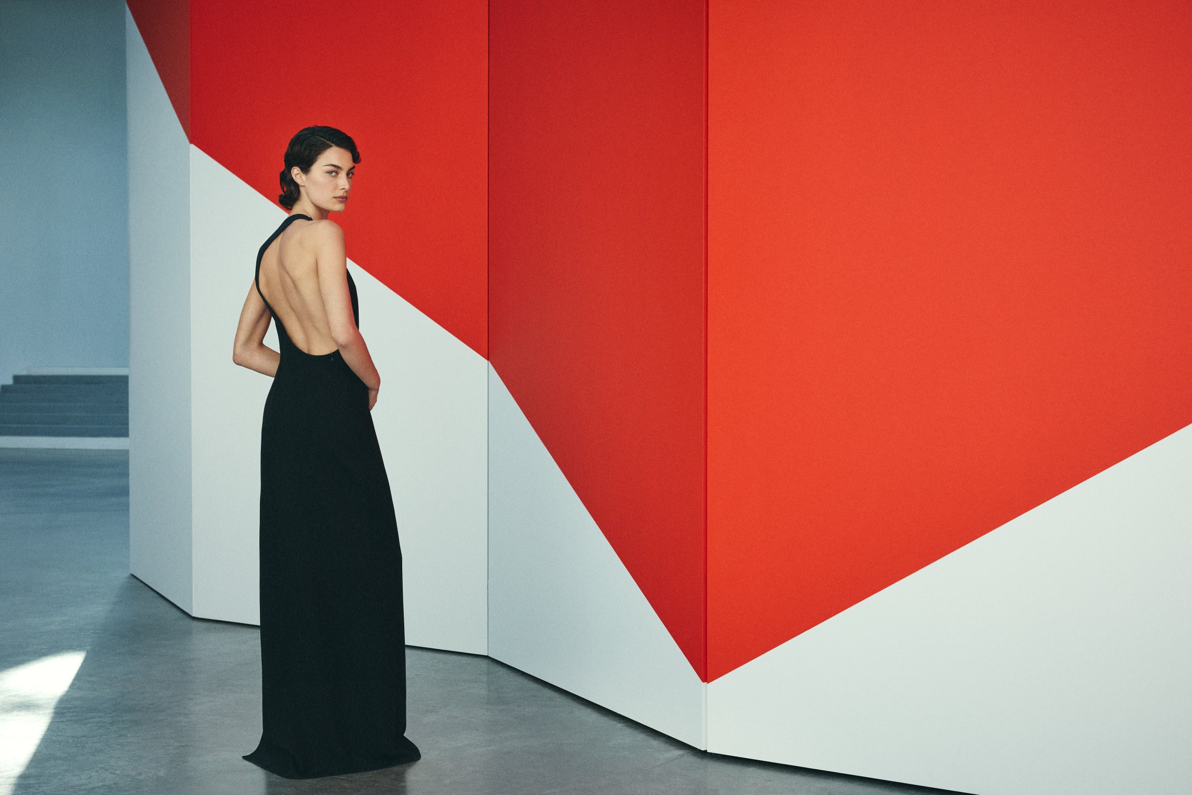 A model stands in three-quarter profile wearing a floor-length black halter gown with a deep open back. She glances over her shoulder against a minimalist gallery-like backdrop featuring bold geometric shapes in red and white.