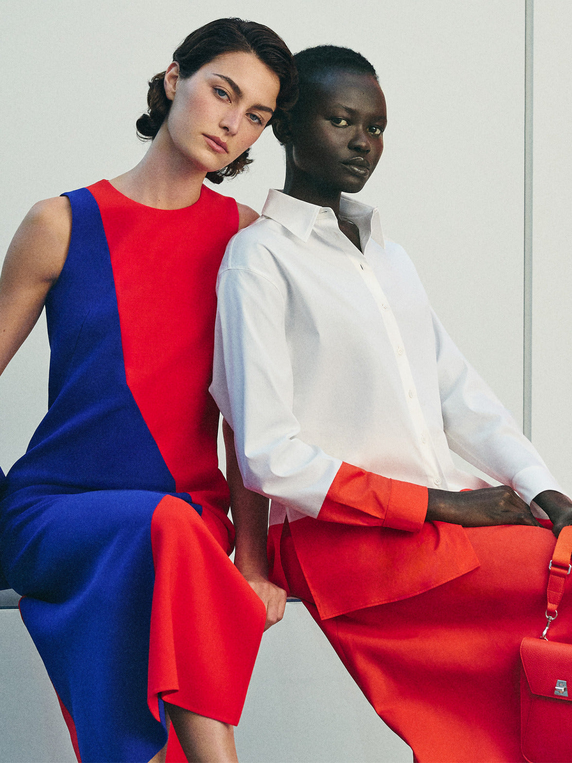 Detailed product shot of a sleeveless red and blue geometric shift dress paired with a vibrant blue suede tote bag. A matching crisp white button-down cotton shirt with orange details is also featured.