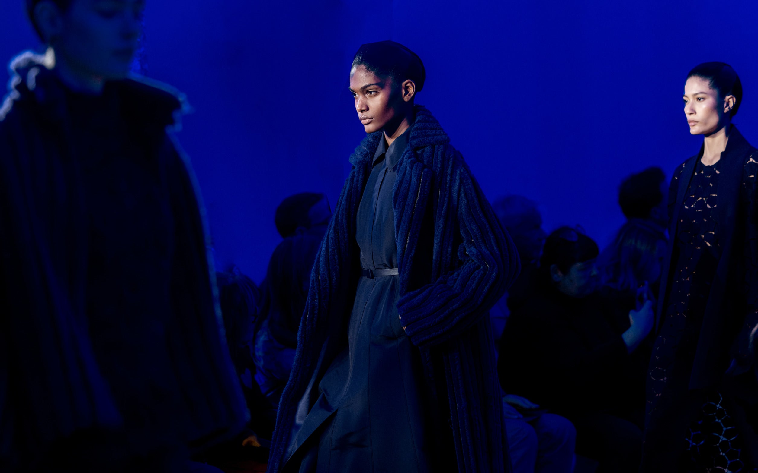 Models on the Akris runway under blue lighting. The central look features a long, textured dark blue ribbed velvet coat layered over a navy dress with a thin leather belt.