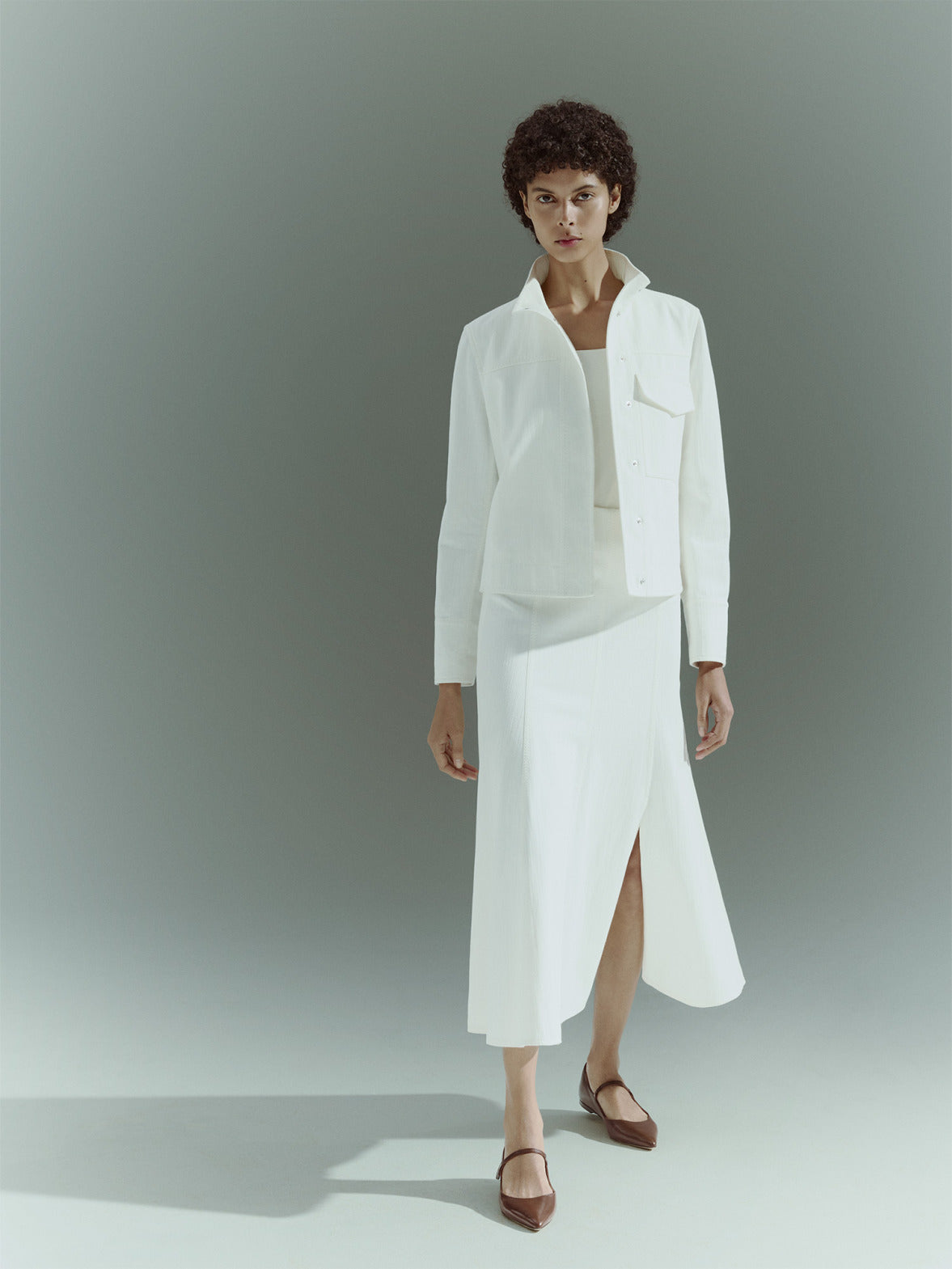 Modernist Akris punto Pre-Fall 2026 editorial look featuring a woman in an all-white textured outfit with a pleated skirt. She wears minimalist black leather pointed-toe Mary Jane flats against a neutral studio backdrop.