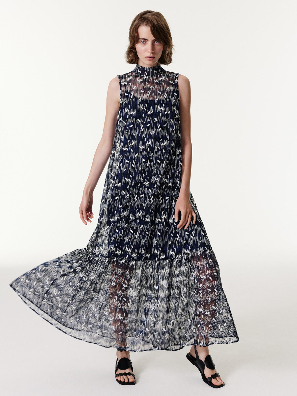 Akris punto Spring 2026 sleeveless midi dress with a high stand collar, featuring intricate geometric embroidery and a pleated, tiered skirt. Crafted from a dark blue technical tulle.